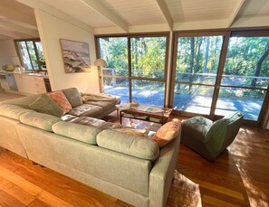 A modern living room with a large sofa, a coffee table, and a view of a balcony with a wooden deck and trees.
