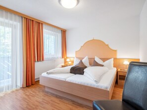 Ein Hotelzimmer mit einem hölzernen Bett, weißer Bettwäsche, einem Holz-Kopfteil, zwei Nachttischlampen, einem Fenster mit transparenten Vorhängen und Blick auf Bäume.