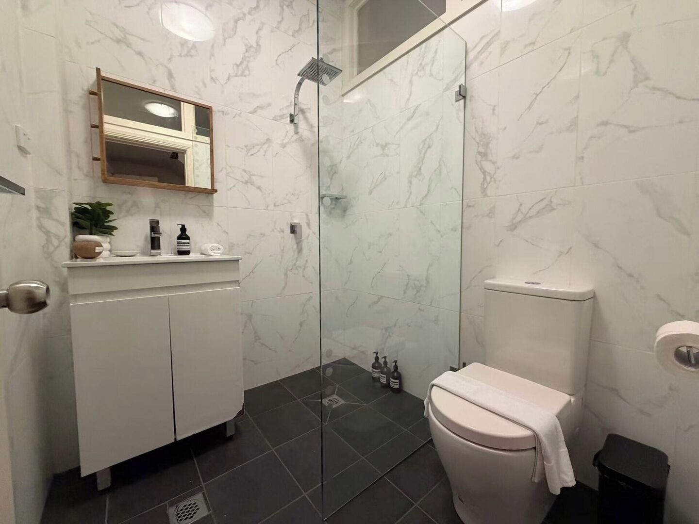 A bathroom with a glass-enclosed shower, a toilet, a sink with toiletries, and a mirror above the sink.