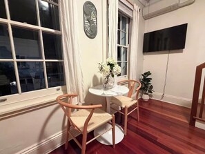 A room with a round table, two chairs, a clock, a potted plant, and a TV mounted on the wall. - KozyGuru | Balmain | Boutique Inner West Stay (Balmain)