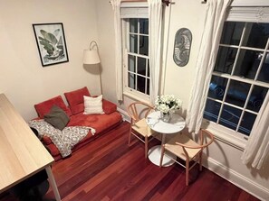 Ein gemütliches Zimmer mit einem roten Sofa, einem kleinen runden Tisch mit Stühlen, einer Lampe und einem Fenster mit Vorhängen.