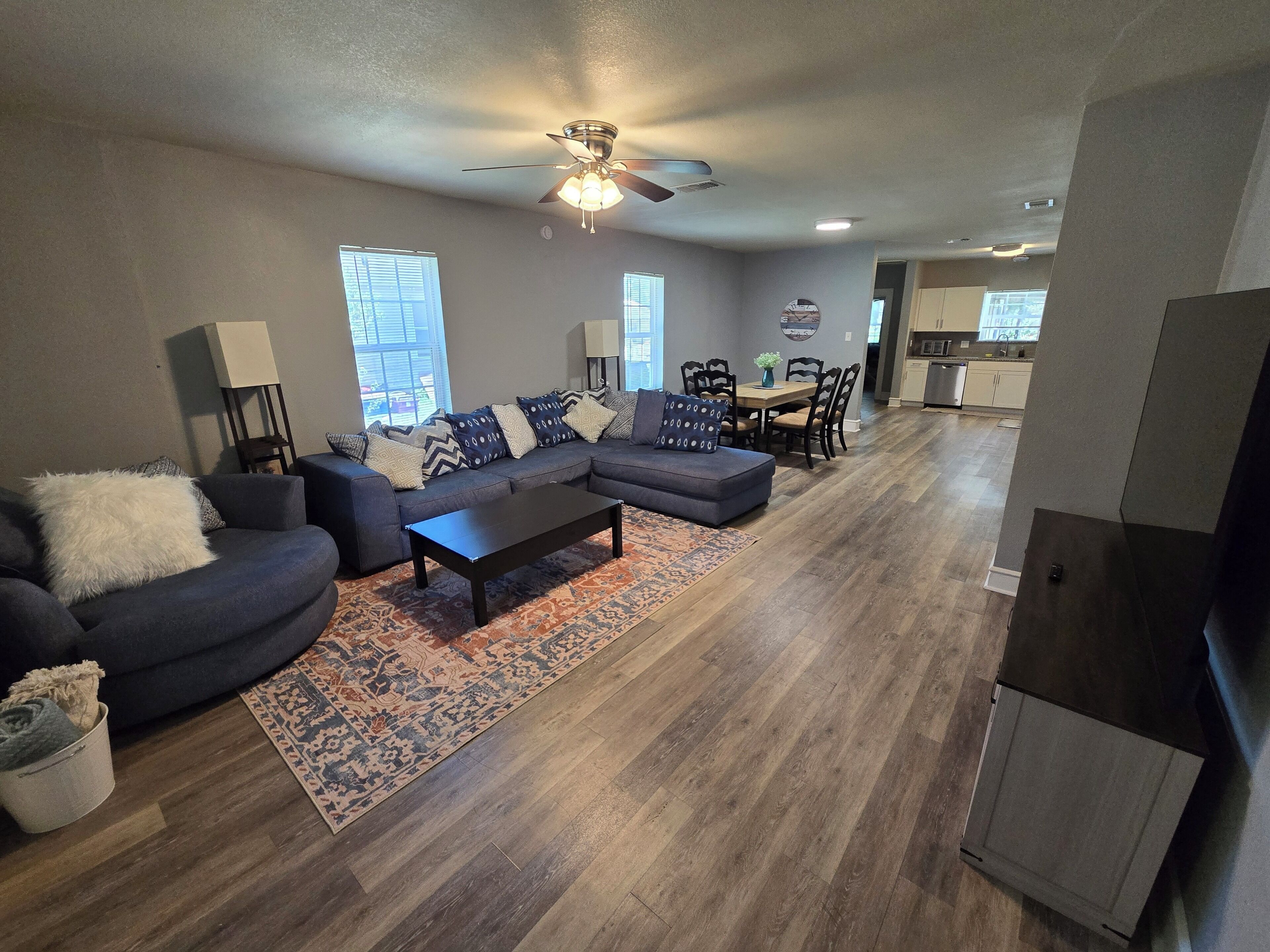 A modern living room with a sectional sofa, a coffee table, and a patterned rug. There is a dining area with a table and chairs, and a kitchen in the background.