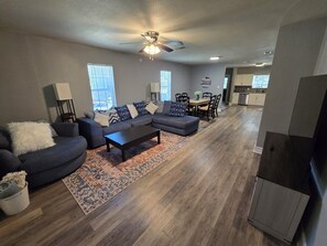 A modern living room with a sectional sofa, a coffee table, and a patterned rug. There is a dining area with a table and chairs, and a kitchen in the background. - The 11th Street Retreat (Temple)