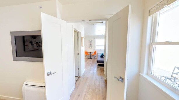 Un pasillo que conduce a una sala de estar con un sofá, sillas, un televisor en la pared y una ventana con vistas.