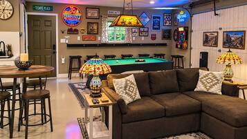 A room with a pool table, a brown sectional sofa, a bar area with a neon sign, and a projector mounted on the ceiling.