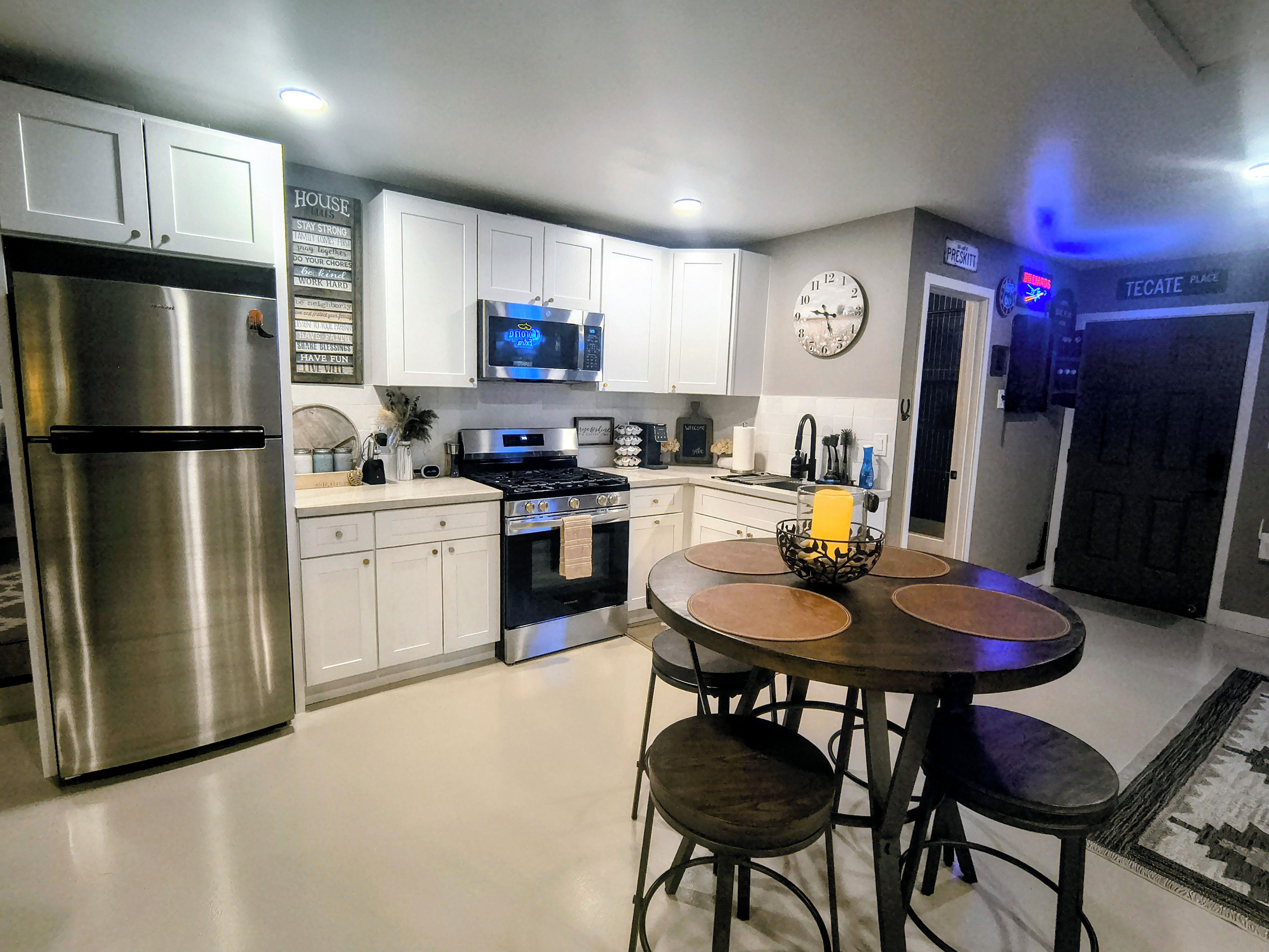 A modern kitchen with stainless steel appliances, white cabinetry, a round dining table with stools, and a clock on the wall.