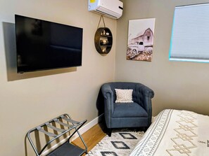 Une pièce dotée d’un téléviseur à écran plat fixé au mur, d’un fauteuil avec un coussin, d’une petite étagère ornée d’objets décoratifs et d’un tableau représentant une grange.