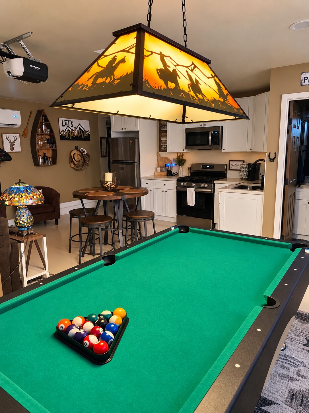Een pooltafel met een steiger voor de witte ballen in een ruimte met een hanglamp, een keukenhoek en barstoelen.