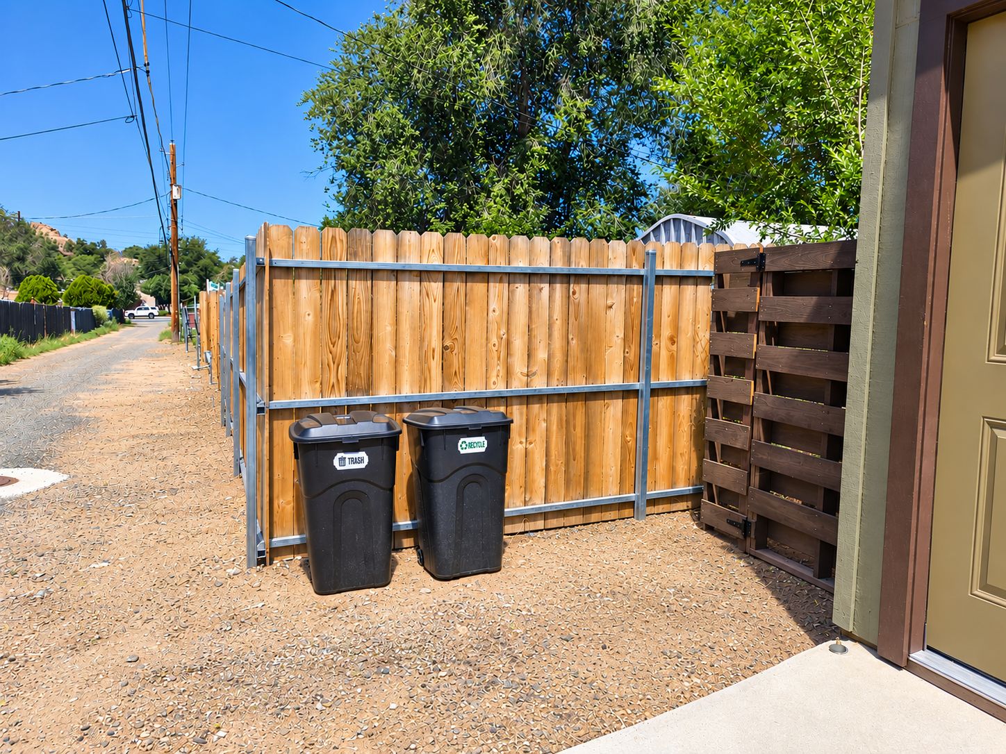 Un área exterior cercada con dos contenedores de basura, uno etiquetado 'TRASH' y otro 'RECYCLE', junto a una valla de madera.