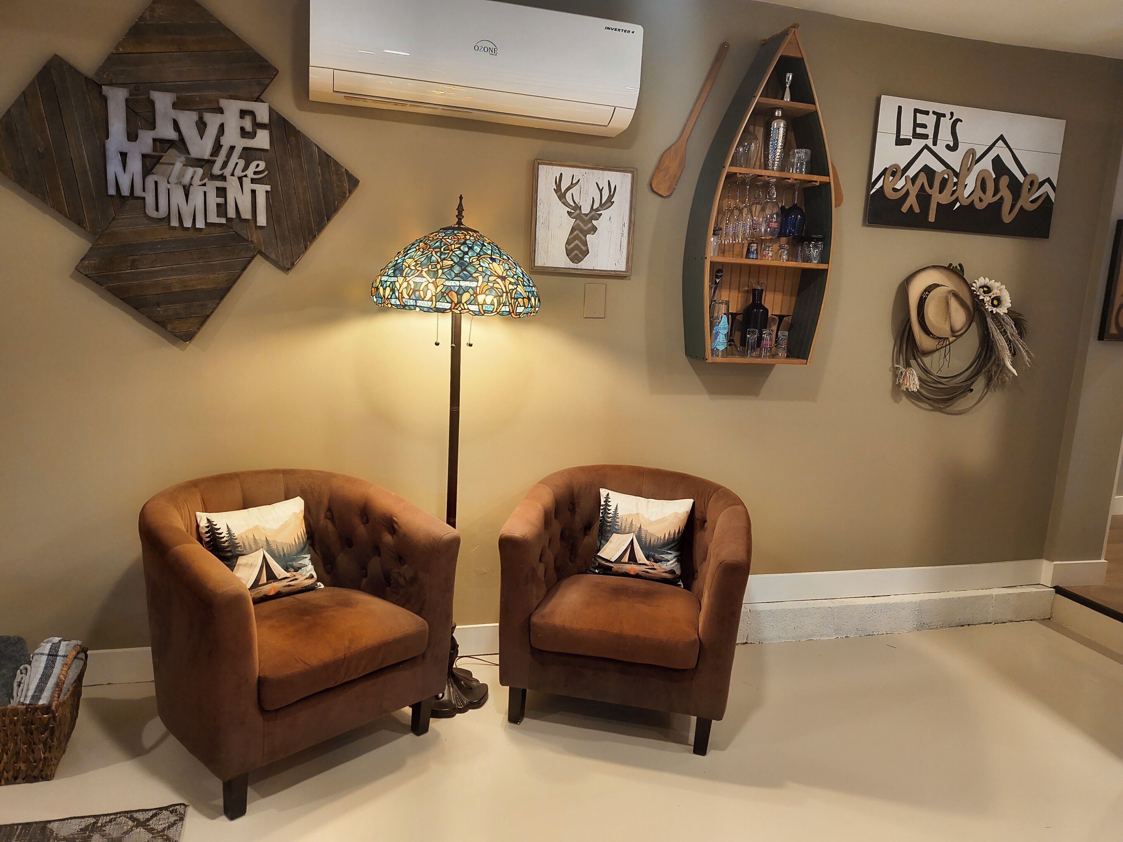 A room with two brown armchairs, a floor lamp, and wall decorations including 'LIVE in the MOMENT' and 'LET'S explore' signs.