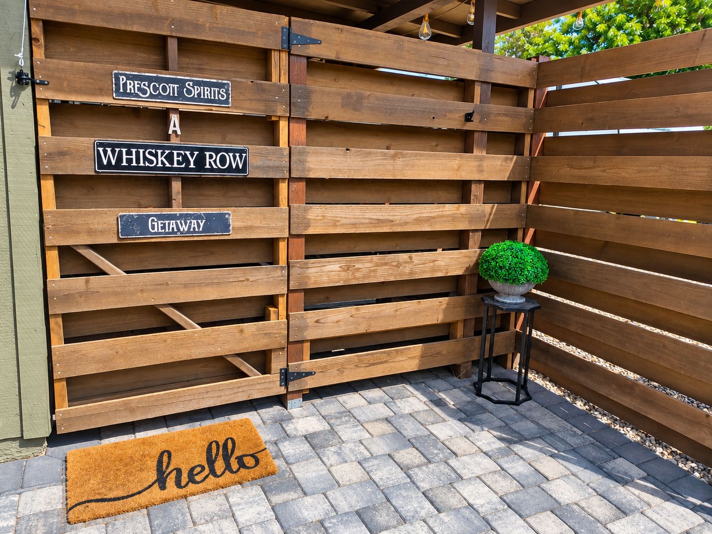 Een houten poort met borden voor 'Prescott Spirits', 'Whiskey Row' en 'Getaway'. Een bloempot en een deurmat met de tekst 'hello' zijn ook zichtbaar.