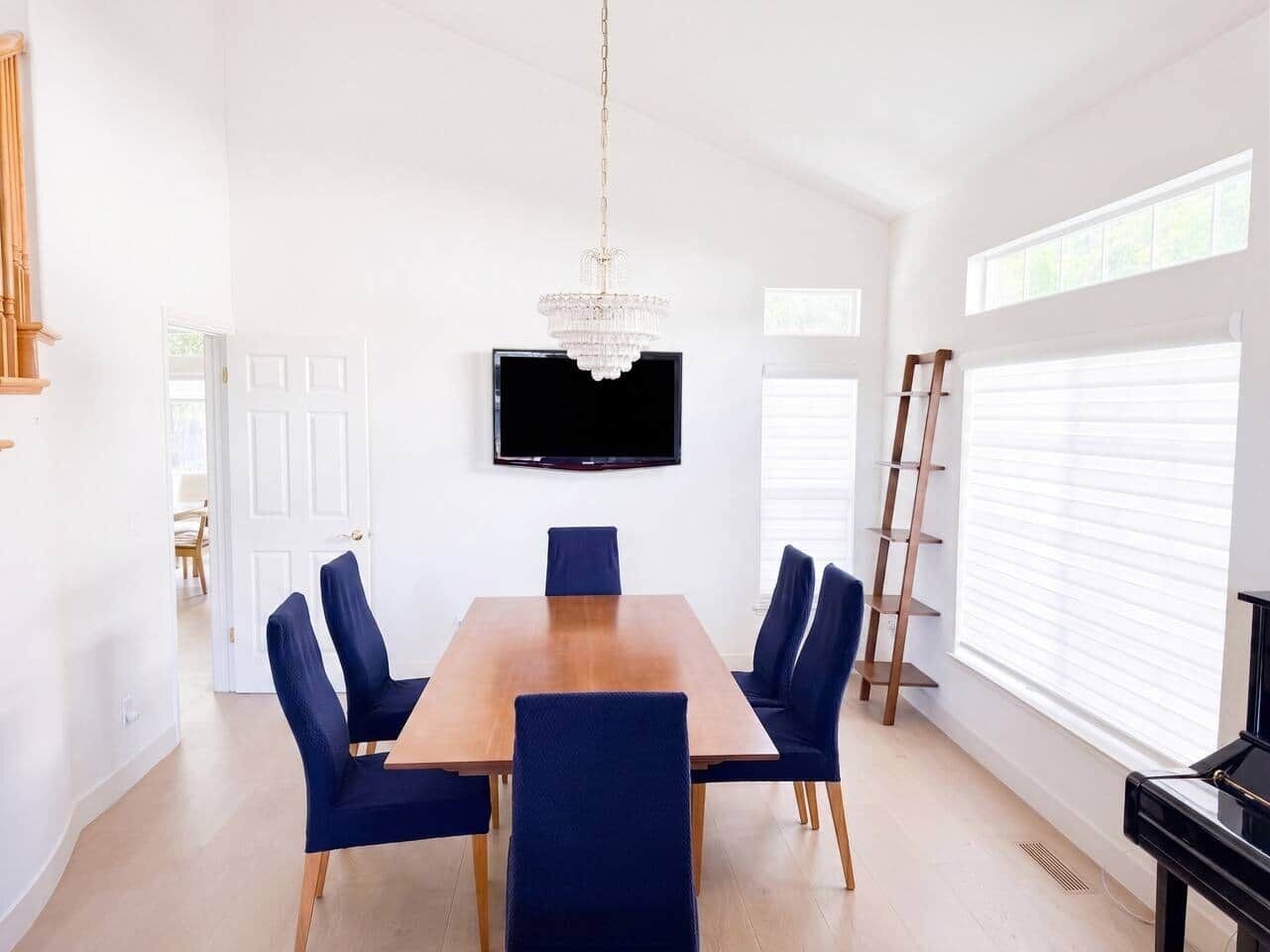 Une salle à manger avec une table en bois et des chaises bleues, un lustre, un téléviseur à écran plat et un piano.