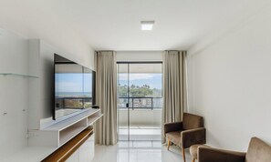 A modern, minimalist living room with a large window, a flat-screen TV, a sofa, and a sliding glass door leading to a balcony. - 2-bedr ap 400 m the sea in a Cond. W/gym Pzo0905 (Ilhéus)