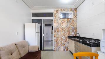 Cocina compacta con refrigerador, estufa y lavamanos. Hay un sofá y una silla de madera en la sala.