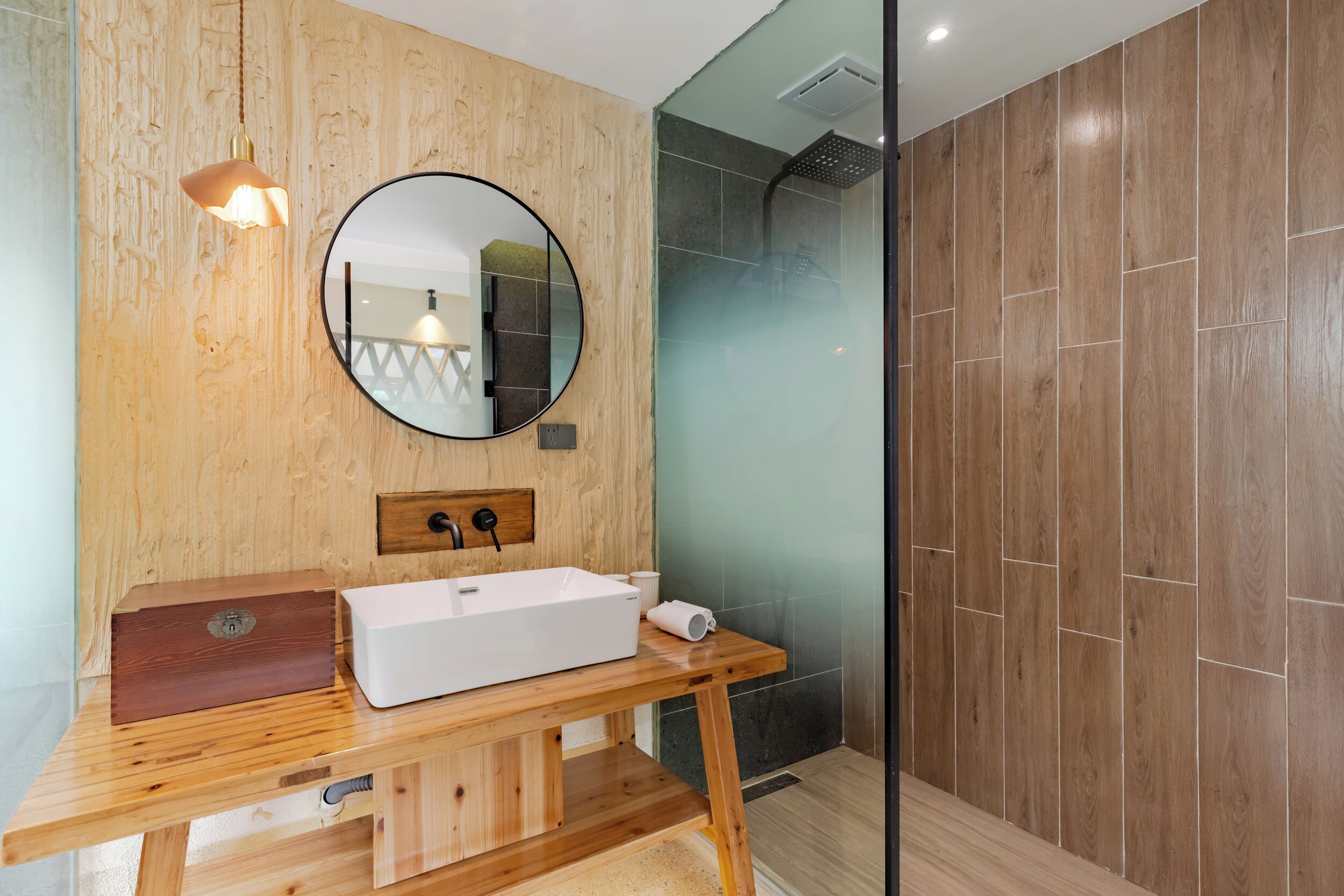 Un baño moderno con tocador de madera, espejo redondo, lavabo blanco y cabina de ducha con mampara de vidrio.