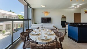 Un comedor moderno con una mesa redonda dispuesta para seis personas, rodeada de sillas de mimbre, una lámpara colgante y una isla de cocina con encimera azul.