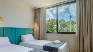 Habitación de hotel con dos camas, cabecera color aguamarina, ventana grande con cortinas y vista a árboles y líneas eléctricas.