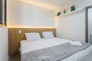 Una cama bien tendida con sábanas blancas, una manta gris a rayas y una toalla blanca al lado. Se observa un cabecero de madera, una estantería blanca con plantas, y una puerta a la derecha.