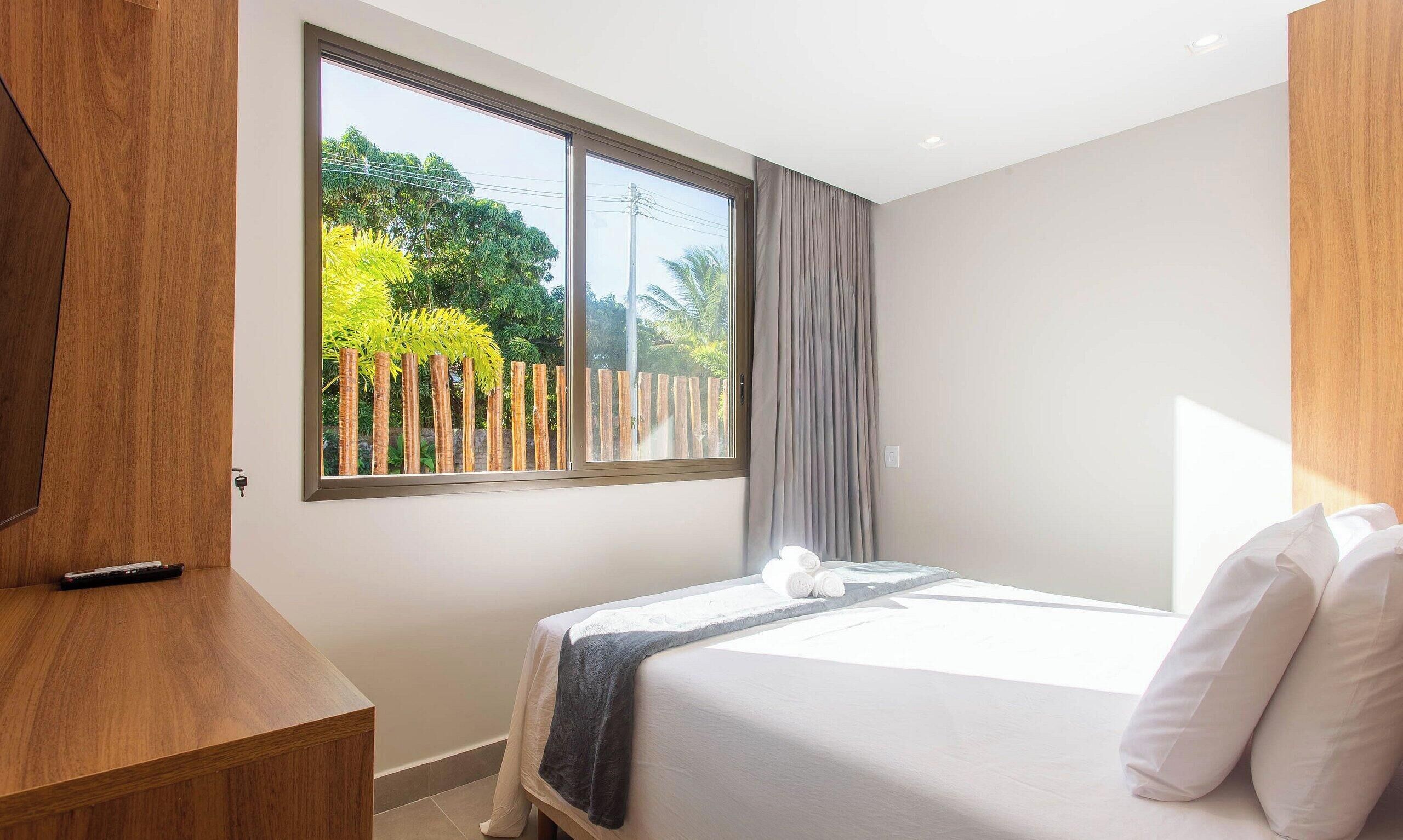 Habitación de hotel con un ventanal grande, una cama con ropa blanca, una mesita de noche de madera y vistas a zonas verdes.
