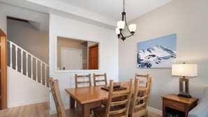 A dining area with a wooden table and chairs, a chandelier, a painting of a mountain, and a lamp on a side table. - Modern Whistler Stay | Full Kitchen & Fireplace for Cozy Nights (Squamish-Lillooet)