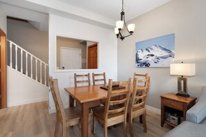 Zona pranzo con tavolo e sedie in legno, un lampadario, un quadro raffigurante una montagna e una lampada su un tavolino.