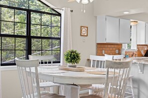 A dining area with a round table, white chairs, a potted plant, and a view of greenery through large windows. - 2BR/2BA Branson condo with king beds, pool, hot tub, and balcony. (Branson)