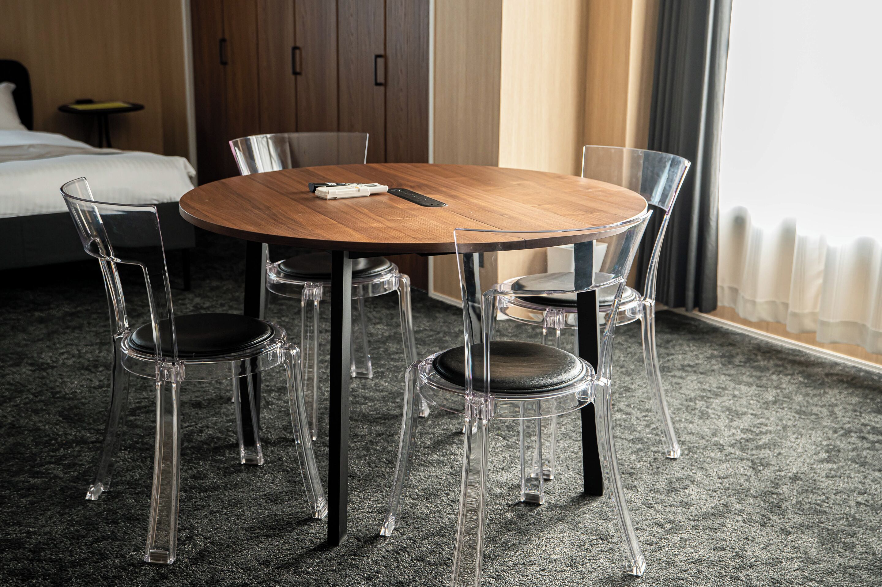 A hotel room with a round wooden table, four clear chairs, a bed, and a window with curtains.