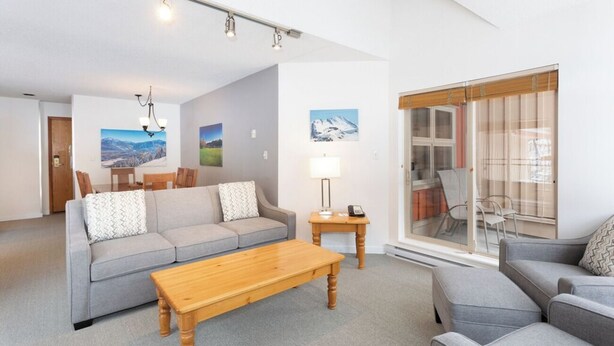 A modern living room with a grey sofa, a wooden coffee table, and a glass door leading to a balcony. - Whistler Escape | Spacious Oasis w/ Mountain Views | Winter Outdoors (Whistler)