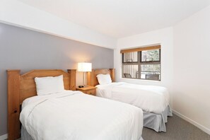 A hotel room with two single beds, a nightstand with a lamp, a window with a view of the outside, and light-colored walls. - Whistler Escape | Spacious Oasis w/ Mountain Views | Winter Outdoors (Whistler)