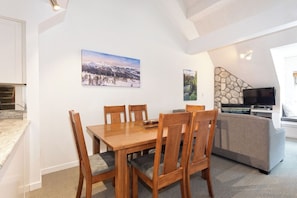 Un coin repas avec une table et des chaises en bois, une cuisine dotée d’un four encastré et un espace de vie comprenant un canapé et un téléviseur.