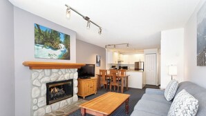 Ein Wohnzimmer mit Kamin aus Stein, einem Fernseher, einem Essbereich und einer Küche im Hintergrund.