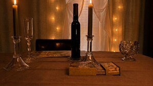 A table with candles, glasses, a bottle, and a box, set against a window with curtains and string lights.