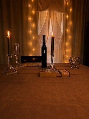 A table with candles, glasses, a bottle, and a box, set against a window with curtains and string lights.