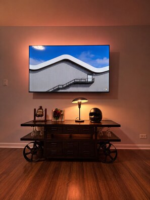 Una habitació moderna amb un gran televisor de pantalla plana penjat a la paret, un escriptori de fusta amb rodes, una làmpada de sobretaula i un casc sobre l'escriptori.