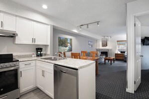 A modern kitchen with white cabinets, a dishwasher, and a stove. There is a dining area with a table and chairs, and a living room with a fireplace. - Whistler Escape | Outdoor Pool & Hot Tub | Creekside Condo (Whistler)