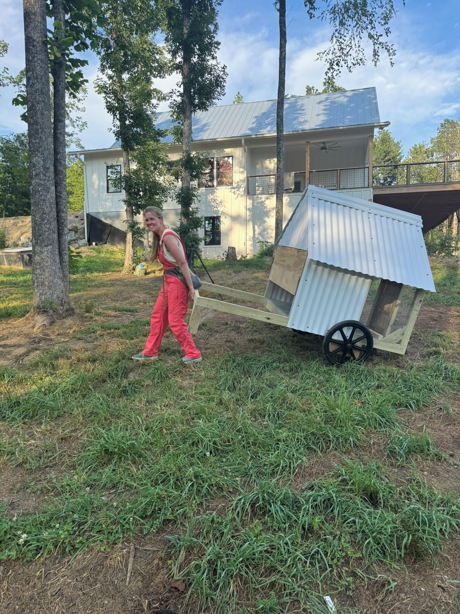 A person carrying a large, white, metal structure with wheels, possibly a portable shelter, in a grassy area with trees and a house in the background.