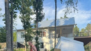 Una persona transporta una estructura metálica blanca grande con ruedas, posiblemente un refugio portátil, en un área con césped, árboles y una casa al fondo.