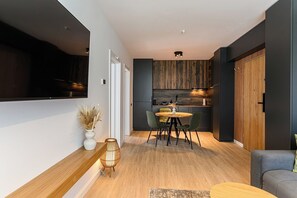 Una cocina moderna con armarios oscuros, un comedor con una mesa redonda y sillas, y una sala de estar con un sofá y un televisor montado en la pared.
