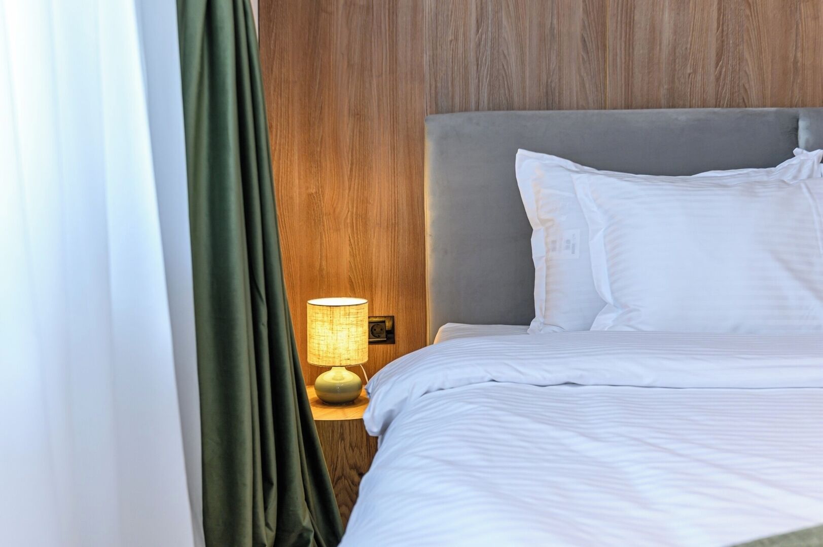 A neatly made bed with white linens, a grey headboard, and a bedside lamp with a yellow shade.