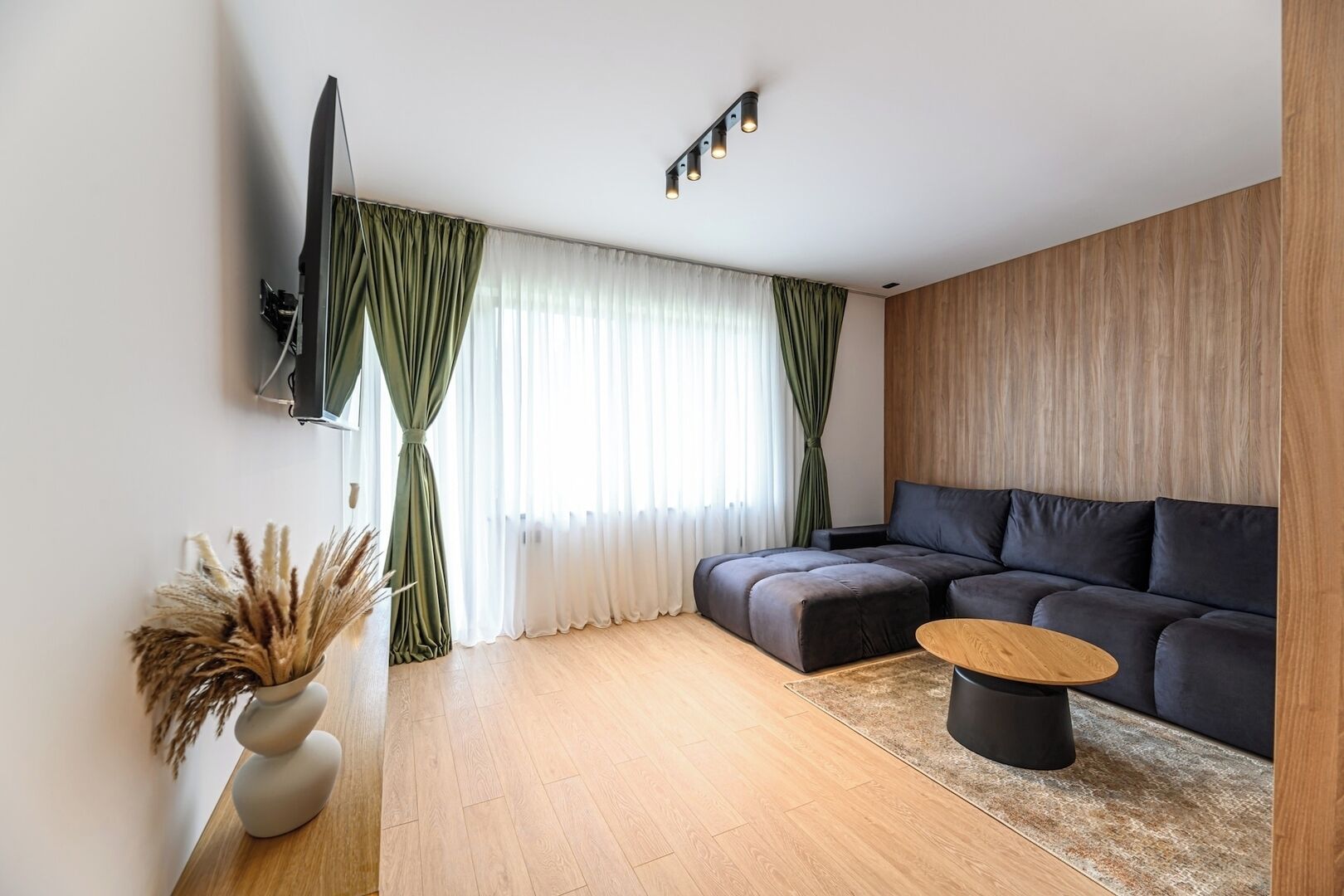 A modern living room with a sofa, a coffee table, a TV mounted on the wall, and a window with curtains.