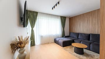 A modern living room with a sofa, a coffee table, a TV mounted on the wall, and a window with curtains.