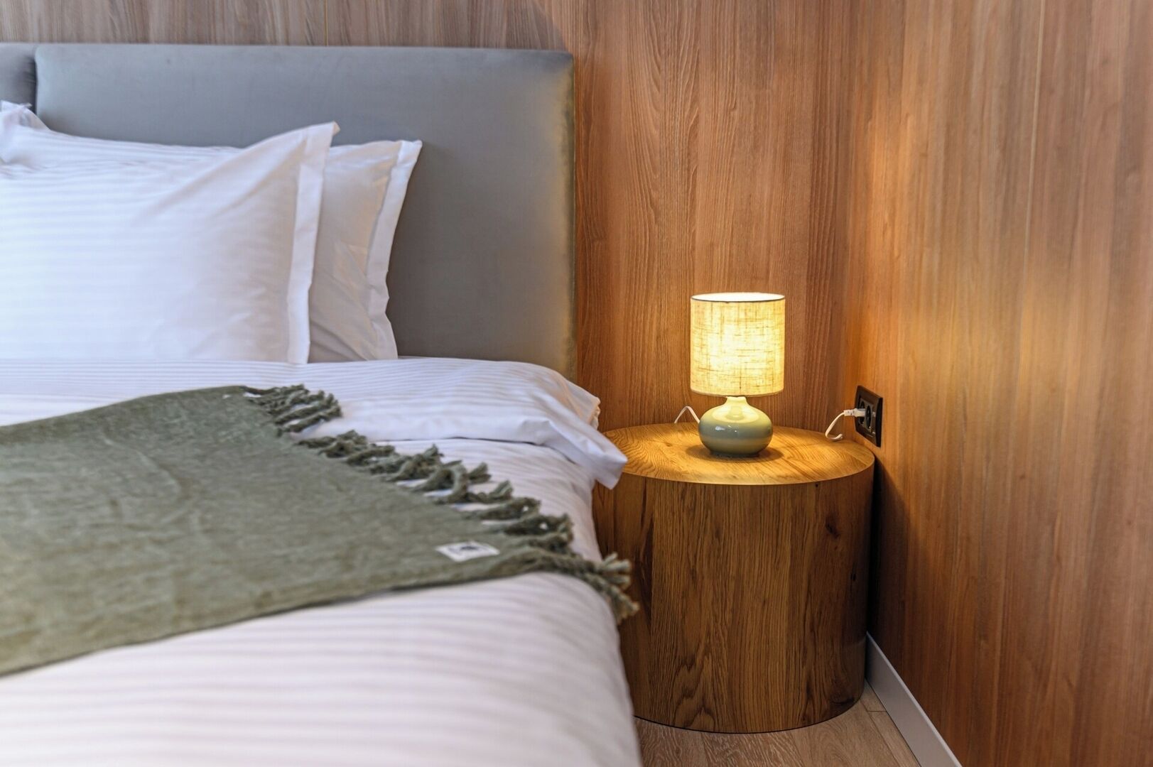 A neatly made bed with white linens and a green throw blanket, a round wooden nightstand with a lamp, and a wooden wall paneling.