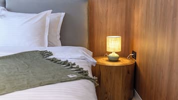 A neatly made bed with white linens and a green throw blanket, a round wooden nightstand with a lamp, and a wooden wall paneling.