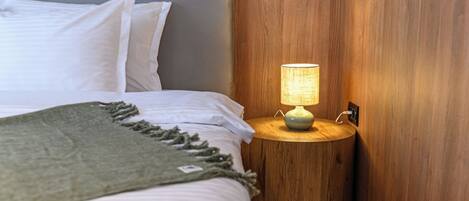 A neatly made bed with white linens and a green throw blanket, a round wooden nightstand with a lamp, and a wooden wall paneling.