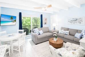 A living room with a grey sofa, white chairs, a round wooden coffee table, and a TV on the wall. - Sanibel Island - Walk to Beach Colony Inn 10 (Sanibel)