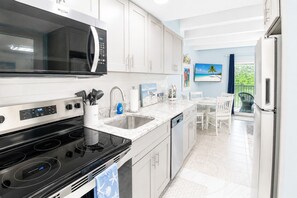 A modern kitchen with white cabinetry, a black stove, a microwave, and a sink. There is a dining area with a table and chairs, and a view of greenery outside. - Sanibel Island - Walk to Beach Colony Inn 10 (Sanibel)