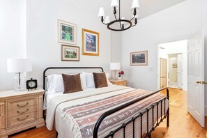 A bedroom with a bed, bedside tables, a chandelier, framed pictures on the wall, and a door leading to another room. - The Oglethorpe Retreat of 1872 | Walk Everywhere (Savannah)