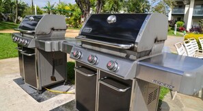 Zwei Genesis tragbare Gasgrills mit Bedienfeld, einer Bürste und einem Schlauch, auf einer Terrasse mit Gartenmöbeln und Pflanzen im Hintergrund.