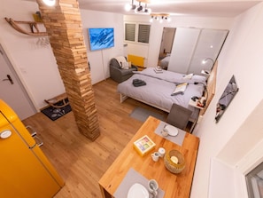 Habitación de hotel pequeña con un pilar de madera, una cama, un escritorio con silla, una mesita con una cesta y un espejo grande.