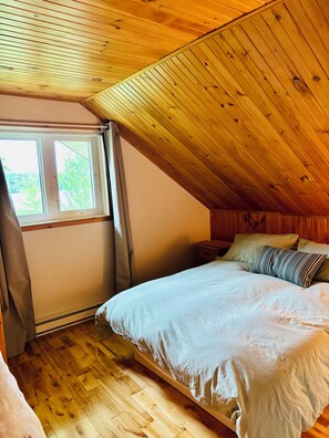 A room with a sloped wooden ceiling, a bed with white bedding, and a window with curtains. - The Secret of the Lake (Rivière-Rouge)